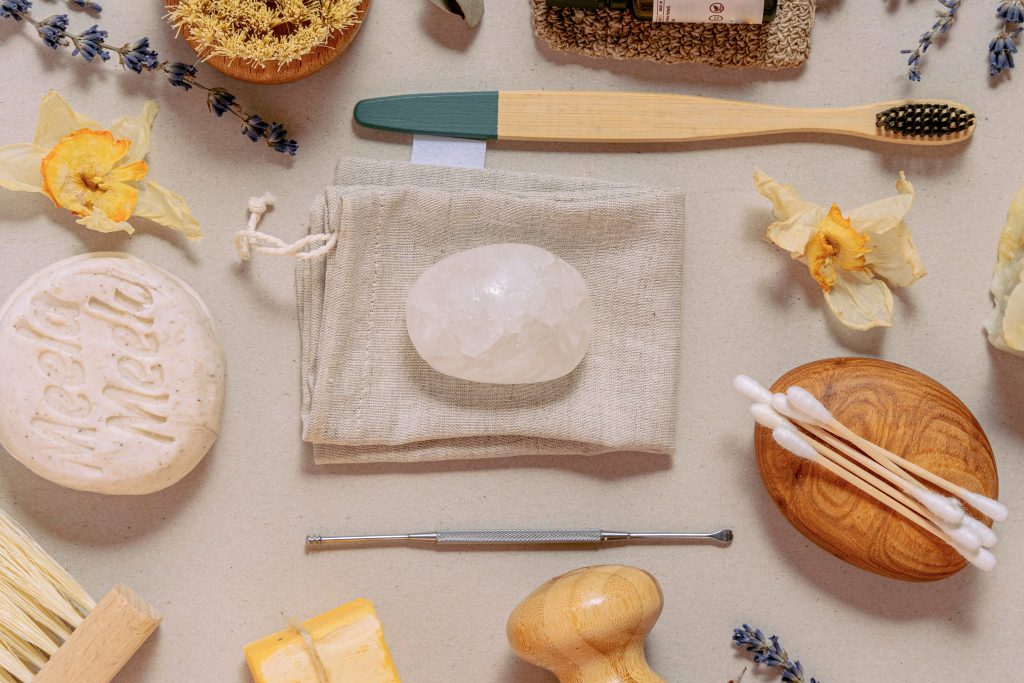 A flat lay of eco-friendly toiletries including a bamboo toothbrush, bar soaps, solid crystal deodorant, reusable cotton swabs, a linen pouch, dried flowers, and other sustainable hygiene items arranged on a neutral surface.