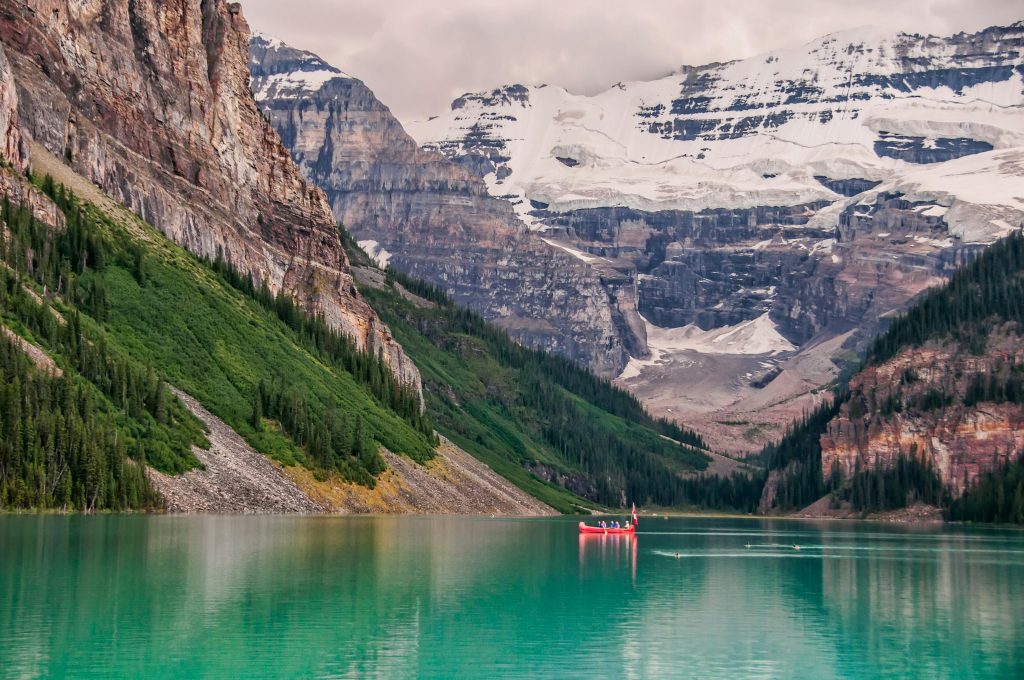 A scenic view of a turquoise glacial lake surrounded by steep rocky cliffs and pine-covered slopes, with a red canoe carrying people floating peacefully on the water below snow-capped mountains.