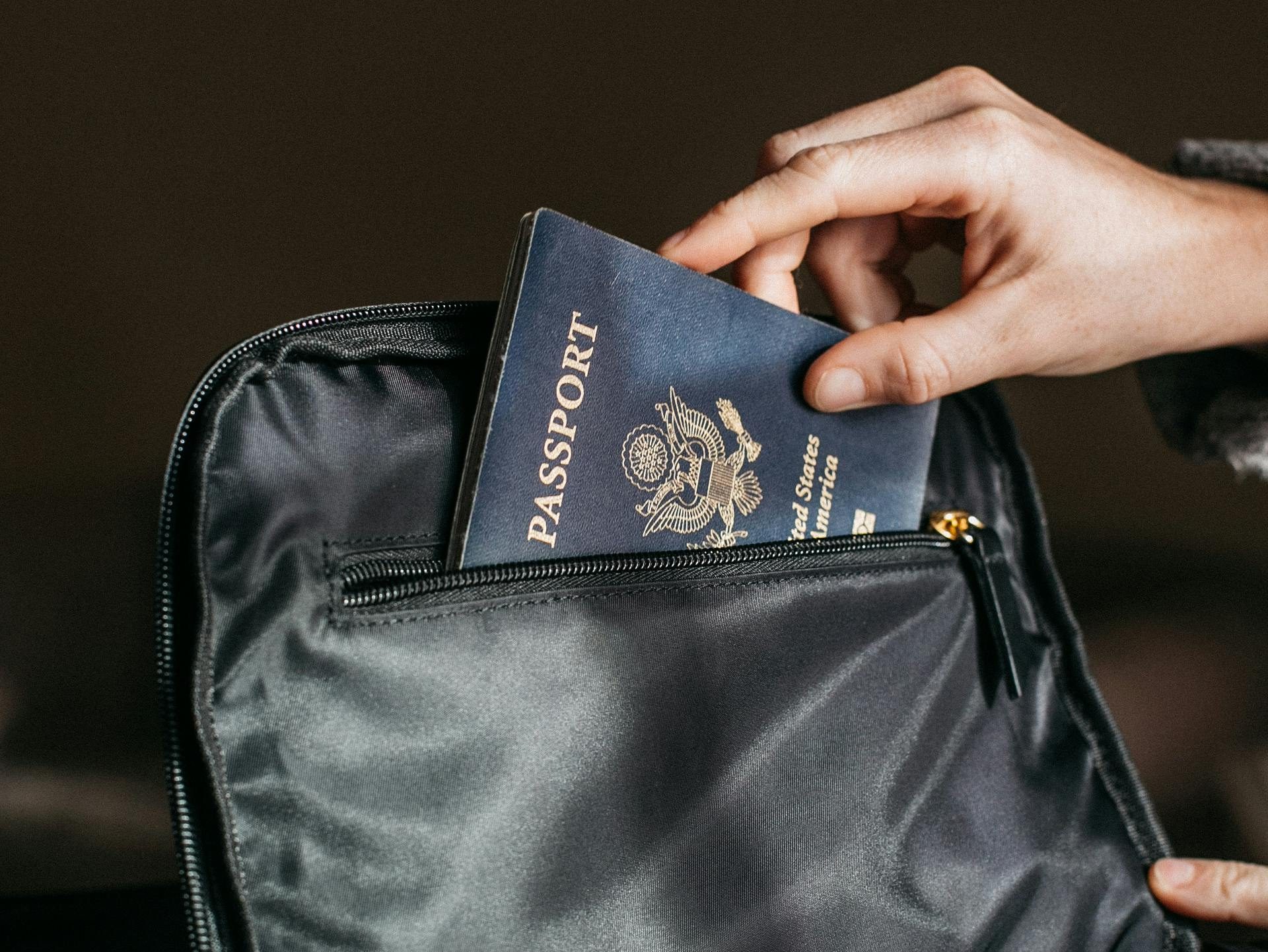A person placing a U.S. passport into the front zippered pocket of a black travel bag.