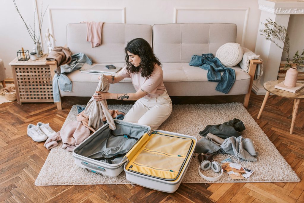 A woman sitting on the floor in a cozy living room, thoughtfully packing clothes into an open suitcase. Travel items like headphones, a camera, and passports are scattered around her, with clothing draped over nearby furniture.