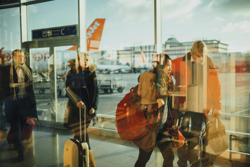 Travelers walk through an airport terminal with suitcases and backpacks, with reflections in the glass and planes visible outside, including a clearly marked EasyJet aircraft.
