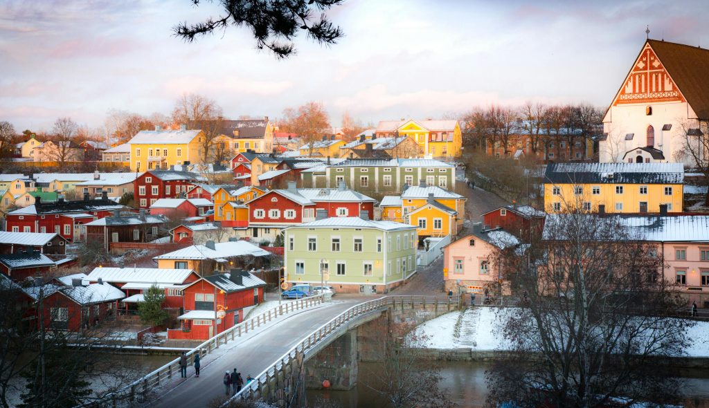 Colorful wooden houses and snow-dusted rooftops in the historic town of Porvoo, Finland, with a pedestrian bridge and white cathedral under soft winter light.
