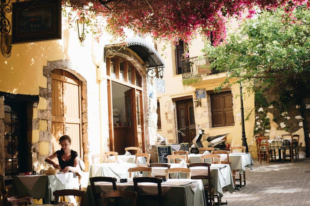 A quiet, sunlit outdoor café in a charming stone alley in Crete, Greece, with a woman setting tables beneath blooming bougainvillea.