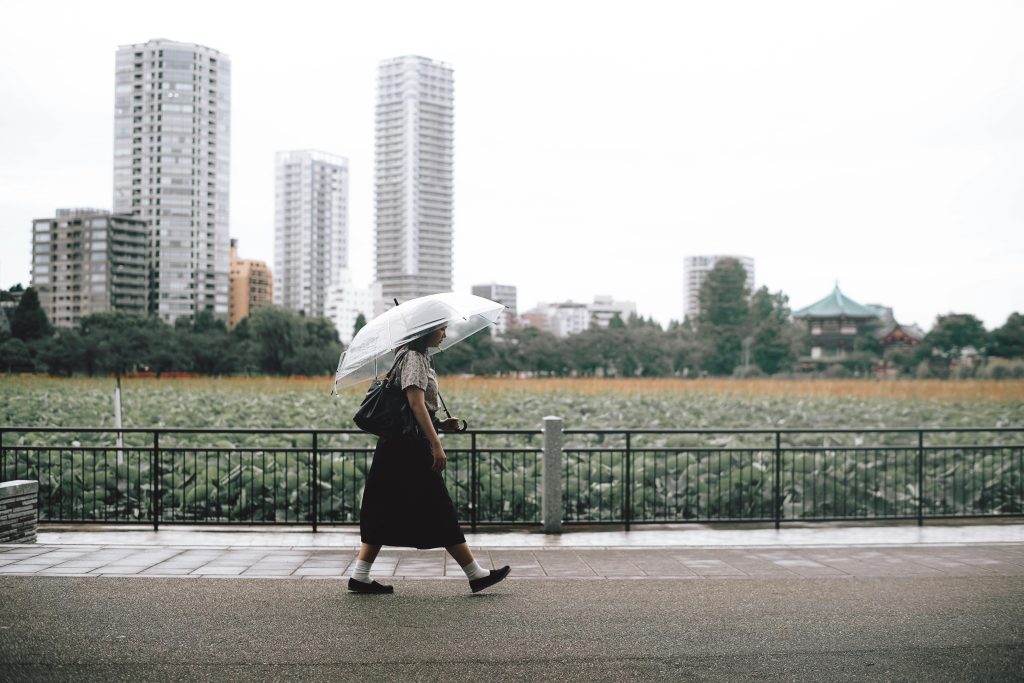 A woman walks along a rainy city path with a transparent umbrella, passing a green park with lotus plants and a skyline of tall modern buildings in the background.