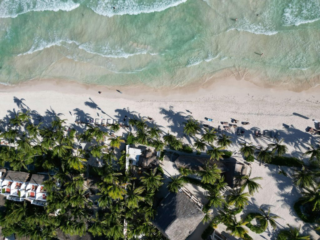 Aerial view of a tropical beach in Riviera Maya, Mexico, with turquoise waves, palm trees, and a beachfront resort lining the white sand.