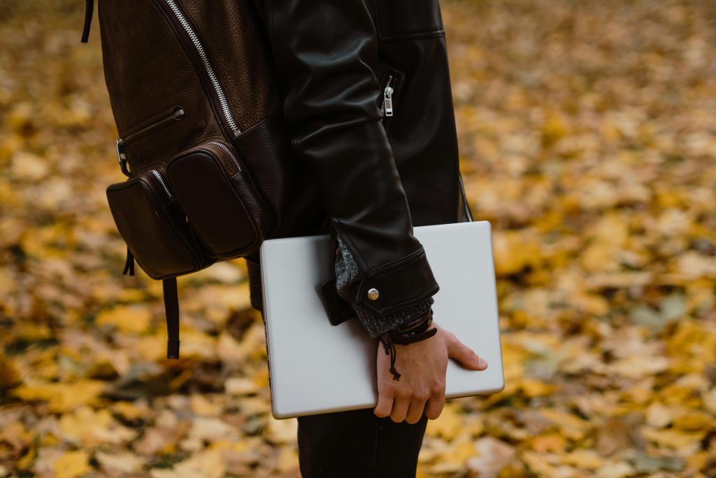 A close-up of a student holding a closed laptop in one hand, wearing a black leather jacket and brown backpack, walking through a park covered in autumn leaves.