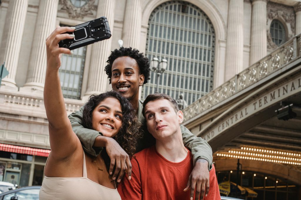Three young friends smile as they take a selfie outside New York City’s Grand Central Terminal, with the historic building’s columns and lights in the background.